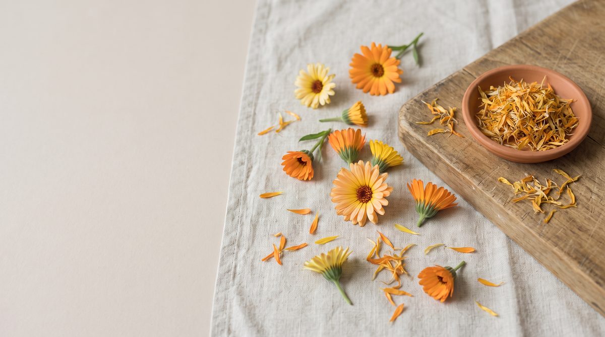 Fiori di calendula freschi e petali essiccati in una ciotola di terracotta su un tagliere di legno e un panno di lino.