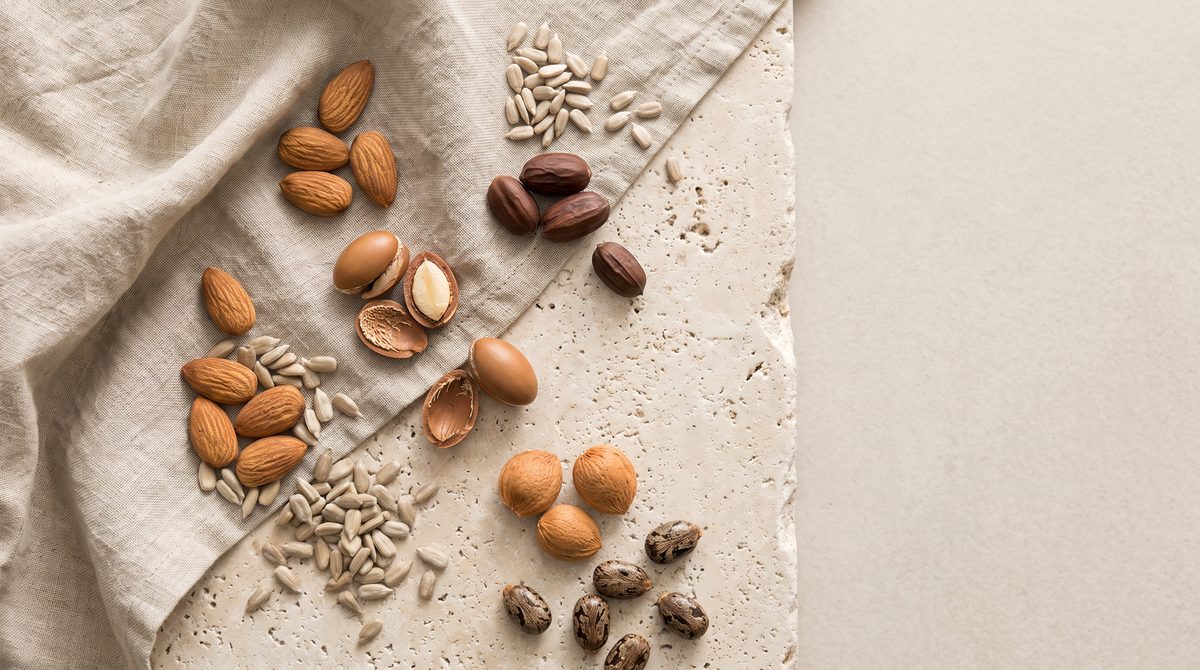 Una composizione dall'alto di mandorle, semi di girasole, noci di argan, noci pecan e semi di ricino su un panno di lino e una superficie di pietra.