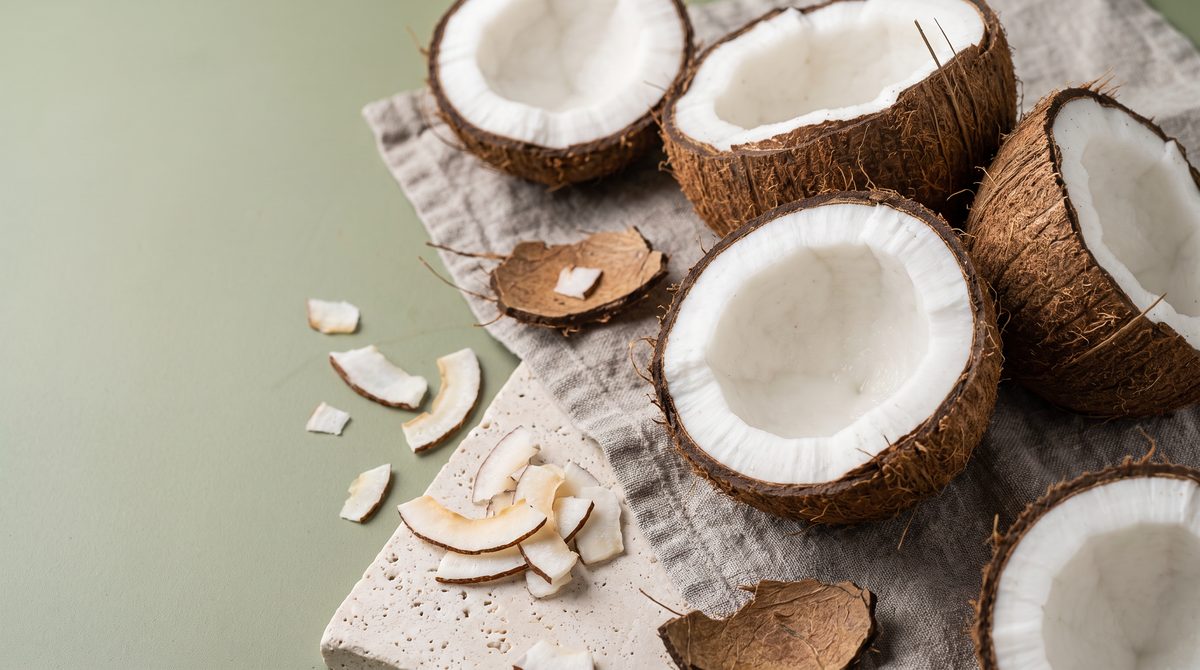 Diverse noci di cocco dimezzate e scaglie di cocco sono disposte su un panno chiaro e una superficie di pietra, su uno sfondo verde chiaro.